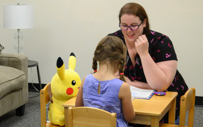 A psychologist interacts with a young patient while seated at a small table with a Pikachu plush seating with them